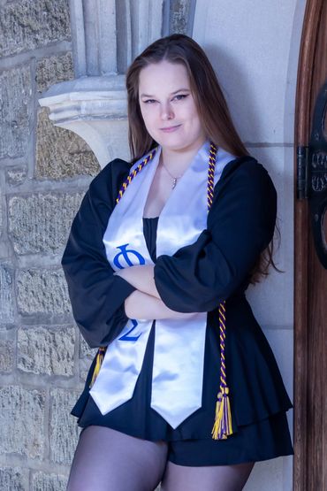 Graduate posing confidently in a black dress with a white stole and honor cords.