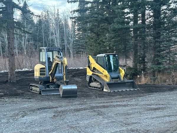 Two yellow construction machines parked on a dirt clearing near trees.