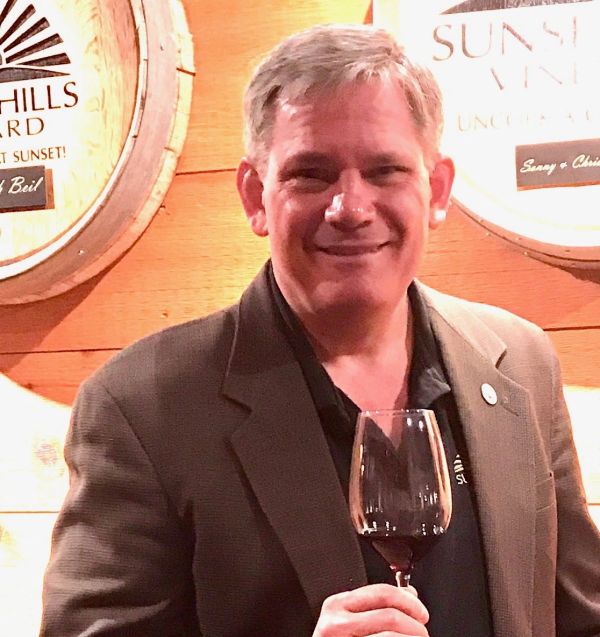 Man smiling with a glass of red wine in a wine cellar.