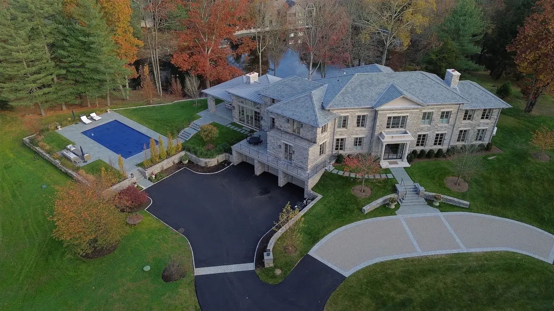 Aerial view of a custom stone home in Greenwich, CT with a slate roof, pool, terraces and a paver-edged driveway beside a pond, built by Jeff Scalise Building Contractor