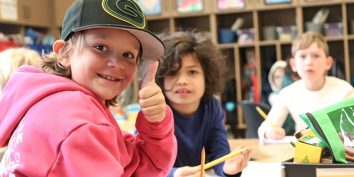 Smiling child in a pink hoodie giving thumbs up in a classroom.