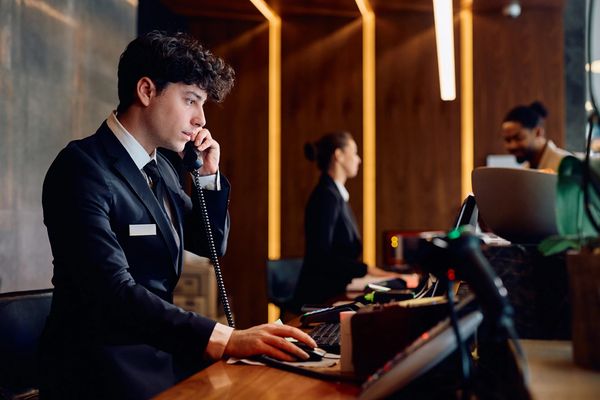 Hotel receptionist answering phone and working on a computer at the front desk.