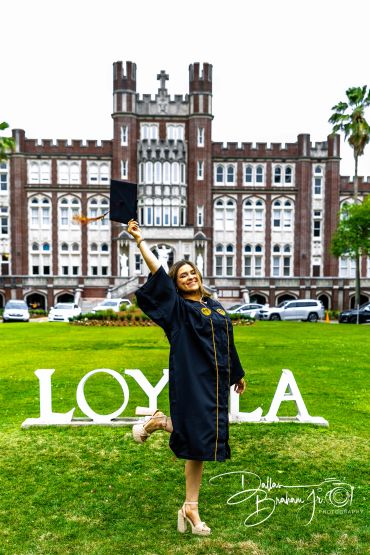 Graduate joyfully celebrates in front of Loyola building, holding cap high.