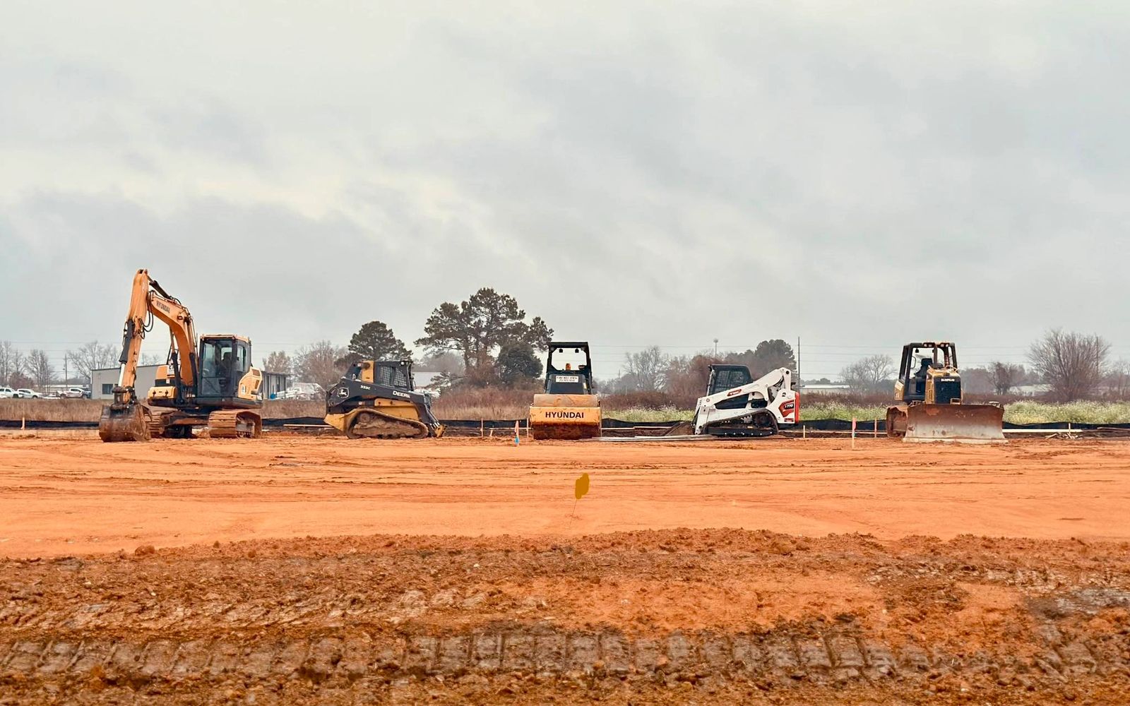 Five construction vehicles lined up on a dirt field under a cloudy sky.