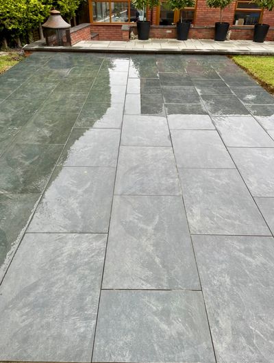 Wet slate patio tiles reflecting greenery and a brick house.