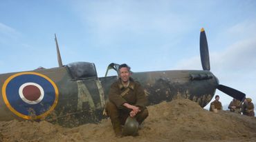 Soldier in vintage uniform sitting by a WWII fighter plane in a sandy area.