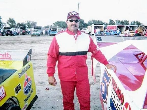 Man in red racing suit stands between two race cars at a track.