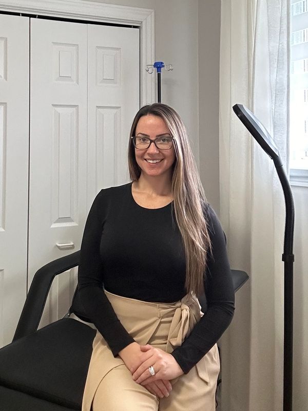 Smiling woman with long hair and glasses sitting on a chair indoors.