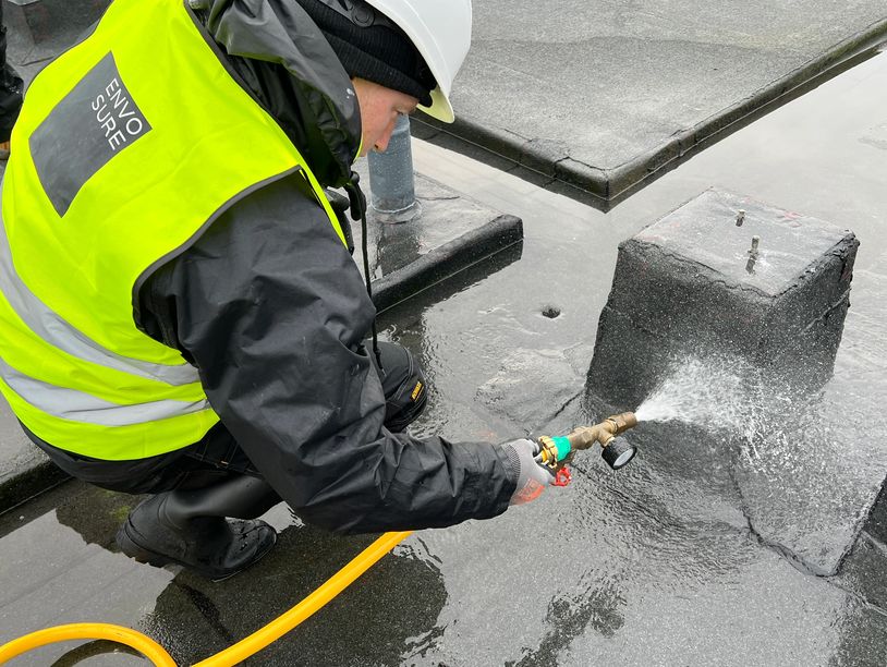 Worker spraying water on a rooftop, wearing a high-visibility vest and hard hat.
