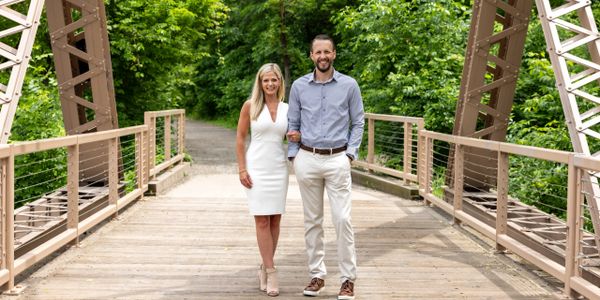 A couple walking arm in arm on a wooden bridge surrounded by greenery.