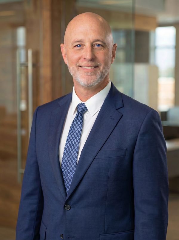 Smiling bald man in blue suit and patterned tie in an office setting.