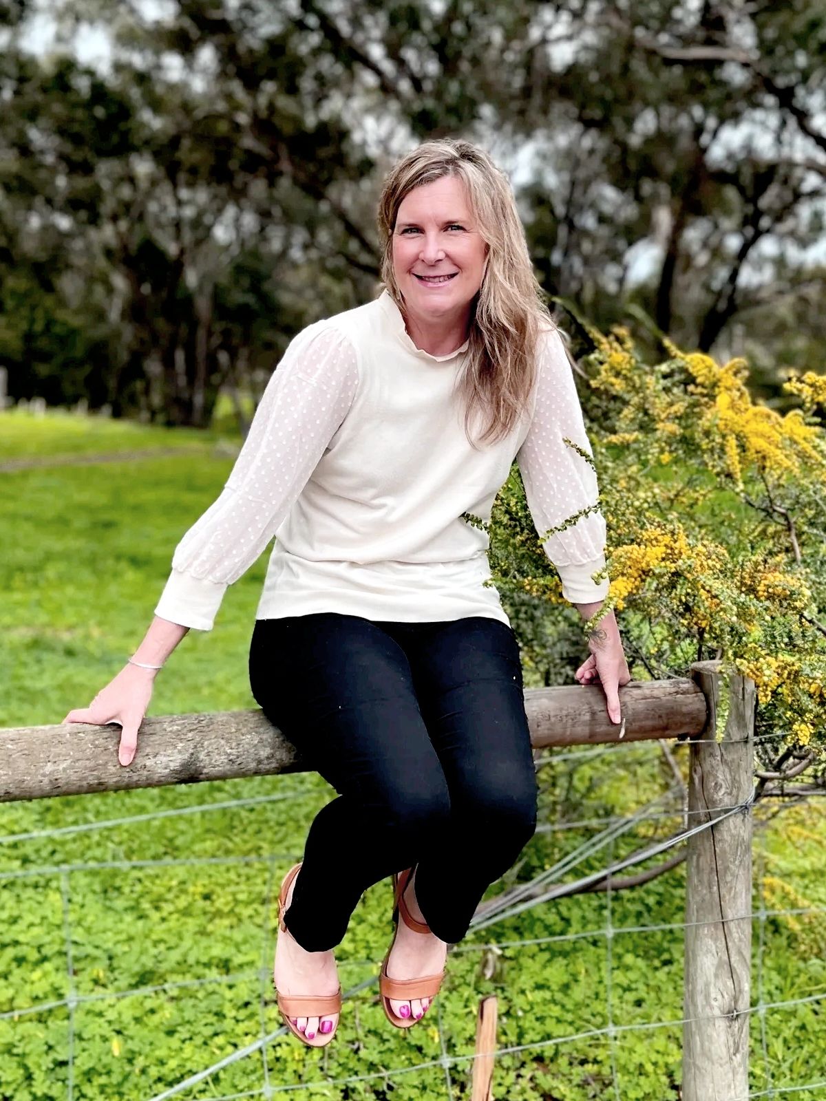 A woman sitting on a wooden fence surrounded by greenery and yellow flowers.