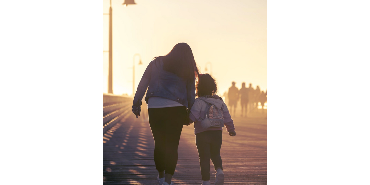 A woman and child walk hand in hand on a sunlit boardwalk.
