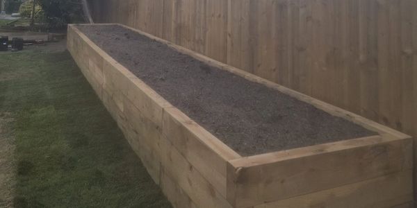 A long wooden raised garden bed filled with soil beside a wooden fence.