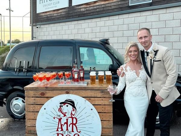 Newlyweds pose beside a rustic bar setup with drinks and a black car.