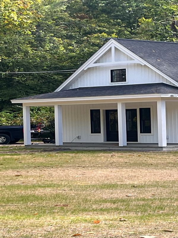 A white house with a black roof and porch in a grassy area near trees.