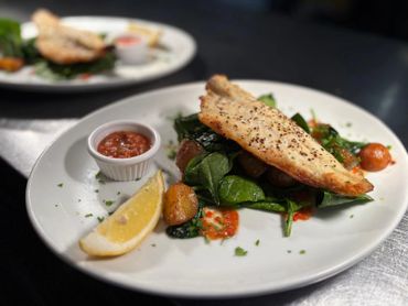 Grilled fish fillet with sautéed spinach, cherry tomatoes, lemon wedge, and dipping sauce on a white plate.