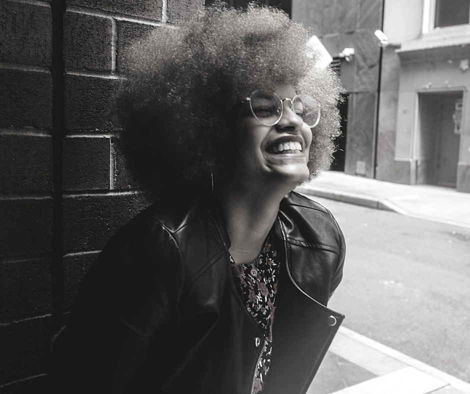 Joyful woman with glasses and afro laughing outdoors against a brick wall.