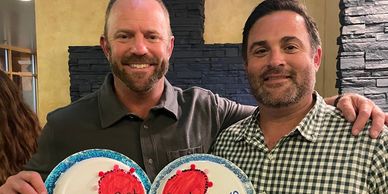 Two men smiling, holding round cakes with red heart designs.