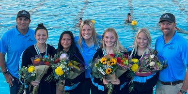 A group of smiling water polo players and coaches holding bouquets by the pool.