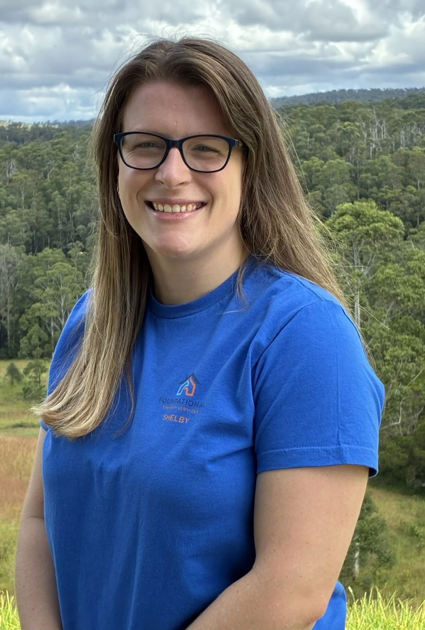 Smiling woman in blue shirt with glasses outdoors, forest background.