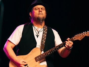 Man passionately playing electric guitar on stage in a black vest and hat.