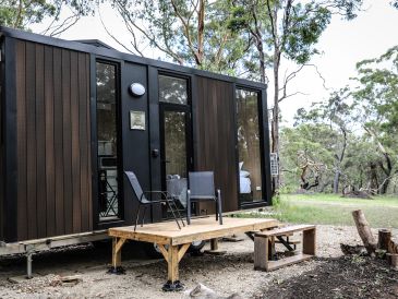 Modern tiny house with wooden deck and chairs in a wooded area.