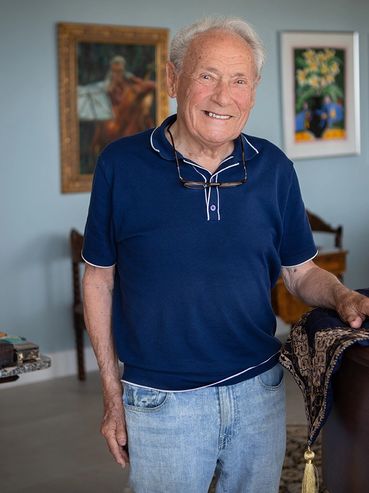 A 99 year old man wearing a blue polo standing next to his piano