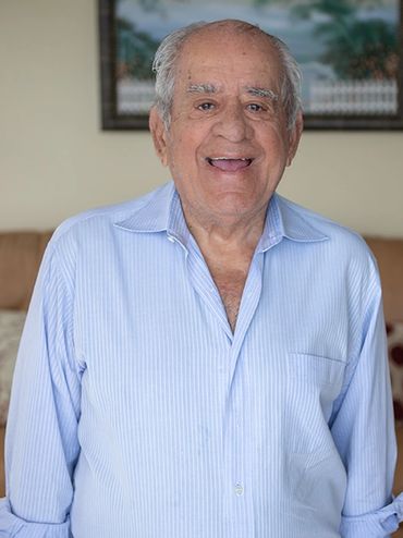 A 96 year old man wearing a pale blue button down shirt smiling.