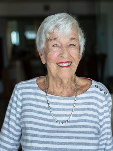 A 102 year old woman smiling with red lipstick wearing a grey and white striped shirt.