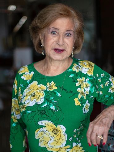 A 96 year old woman with light brown hair and a vibrant green floral shirt on.