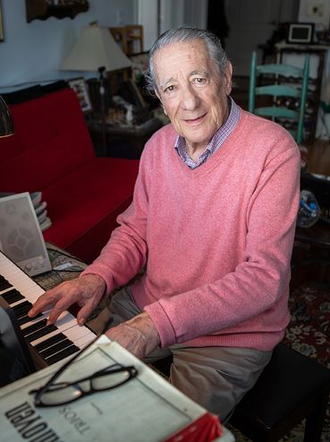A 97 year old man wearing a pink sweater and playing the piano