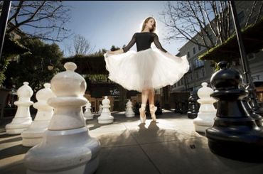 Ballerina dancing on pointe among giant chess pieces outdoors.