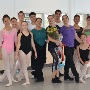 Group of ballet dancers posing in a dance studio, one holding flowers.