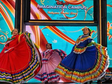 Three dancers in colorful traditional dresses performing on stage at Disney Imagination Campus.