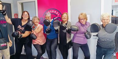 Six senior women wearing boxing gloves and mitts pose energetically together indoors.