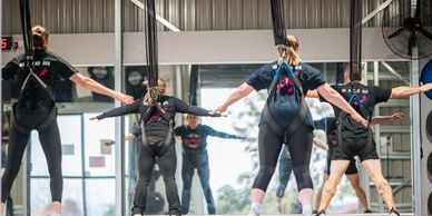 People exercising with harnesses in a gym, stretching arms wide.