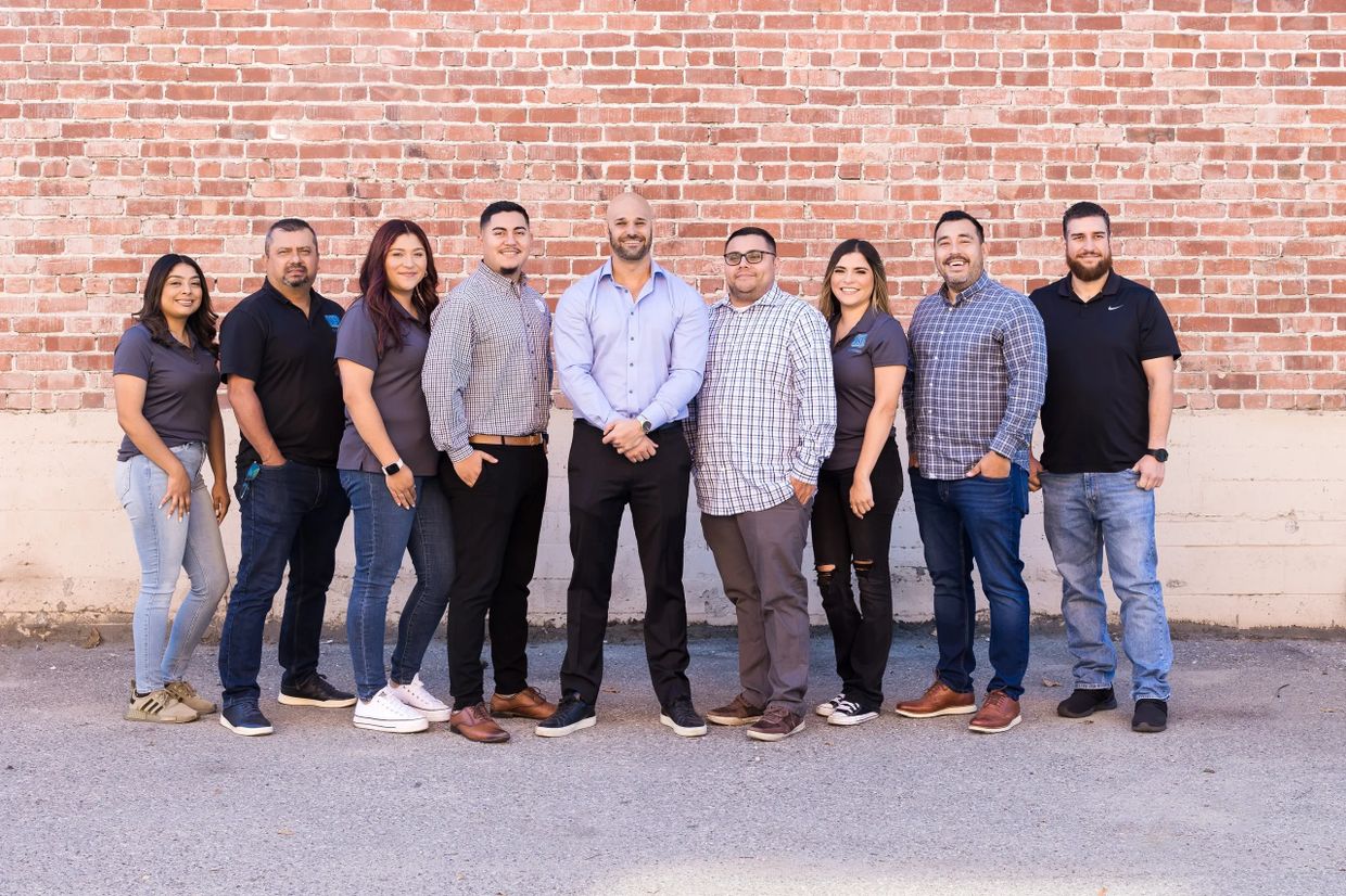 A diverse group of nine people standing against a brick wall, smiling.