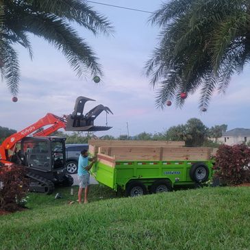 Man working with a mini excavator and trailer between two decorated palm trees.