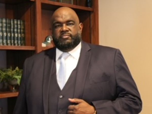 A man in a dark suit and white tie stands confidently indoors.