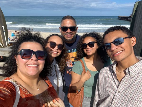 A smiling family of five wearing sunglasses at a sunny beach.
