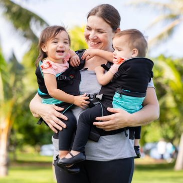 Mom holding her twins in the Front Carry Position.  She is smiling and one of her daughters is laugh