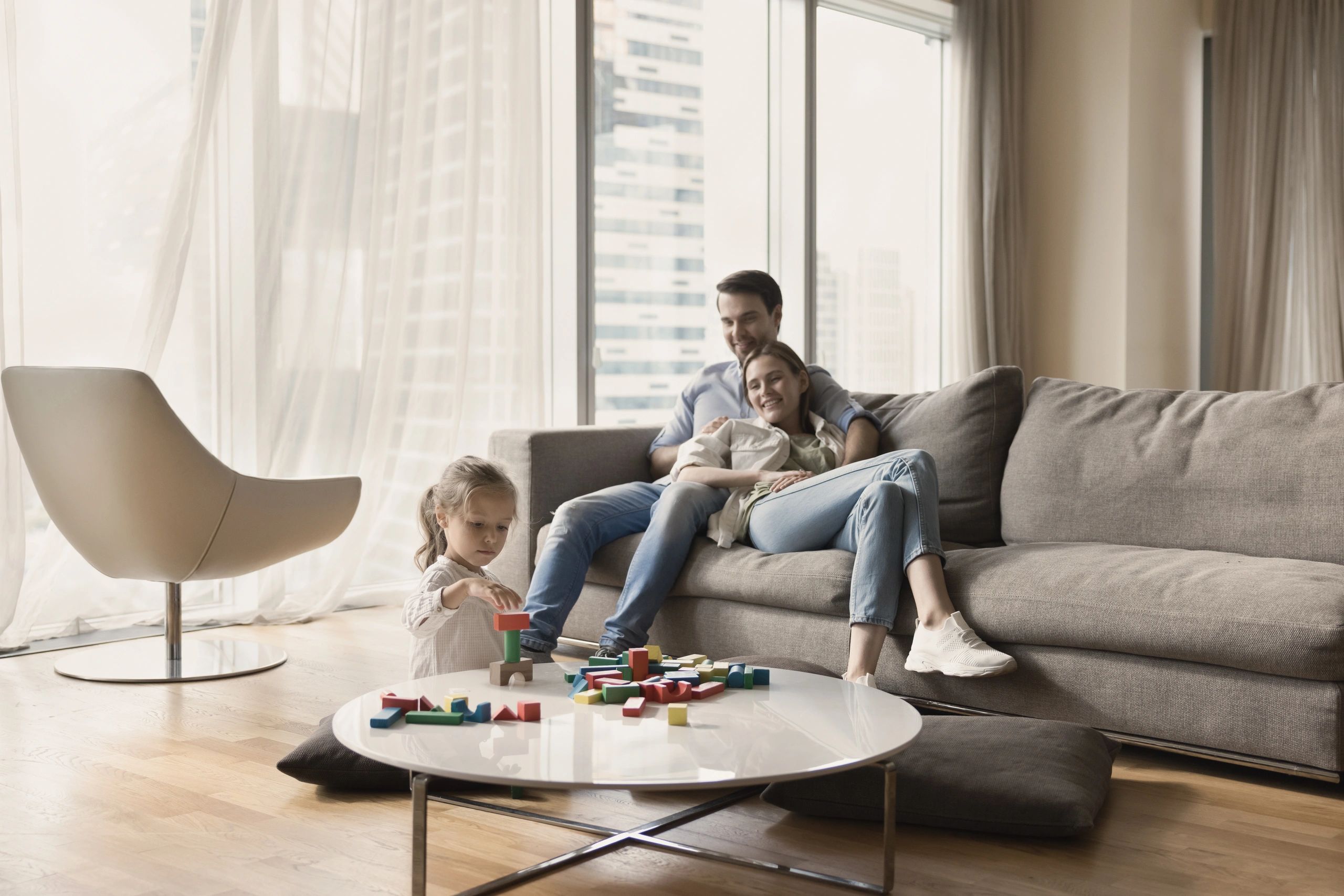 Family in their apartment. Parents sitting on the couch watching daughter play with blocks