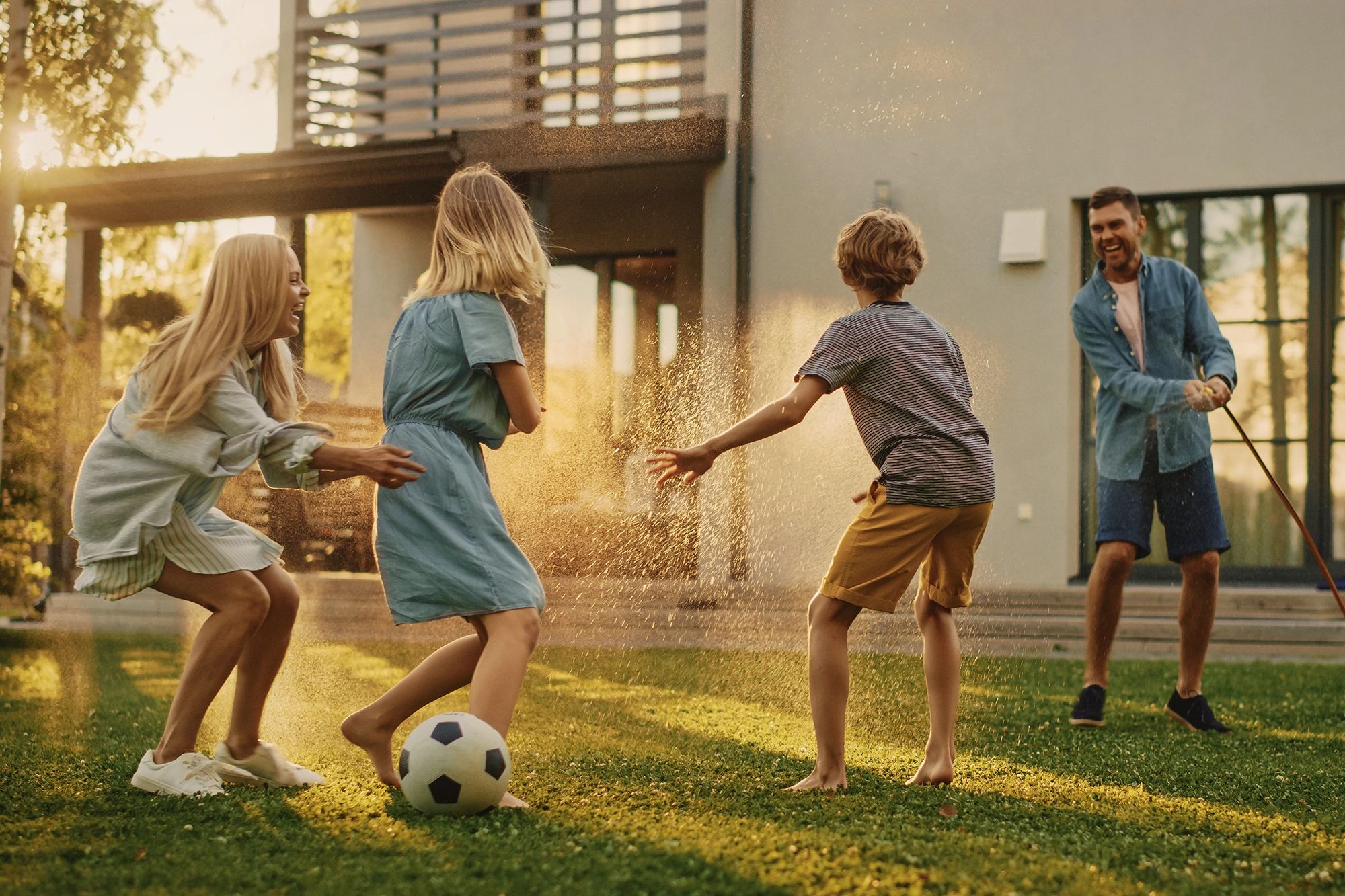 Family of four playing in the backyard of a home. Father has a hose and spraying wife and children.