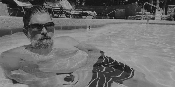 Man relaxing in a swimming pool with sunglasses and striped swim shorts.