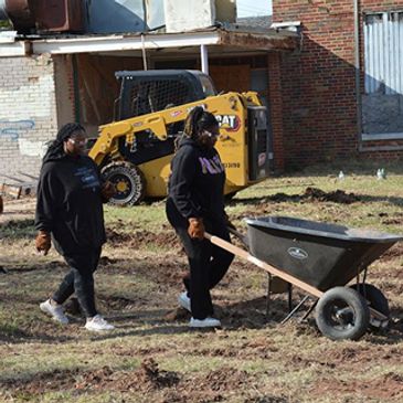 Two people walking with a wheelbarrow on a dirt patch.