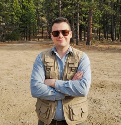 Man in khaki vest and sunglasses in forest clearing.