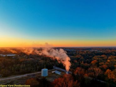 Industrial smoke rising over a forested area at sunset.