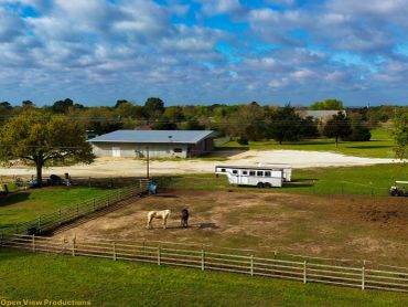 Two horses stand in a fenced paddock near a large barn under a partly cloudy sky.