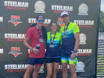 Three triathletes pose with medals in front of Steelman Racing backdrop.